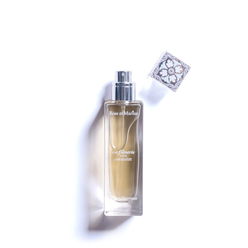 A tall, clear glass bottle of ROSE ET MARIUS’s A Stroll Under the Oratory Eau de Parfum 30ml, unisex with a light golden liquid and decorative floral-patterned square cap, is placed to the side on a white background.