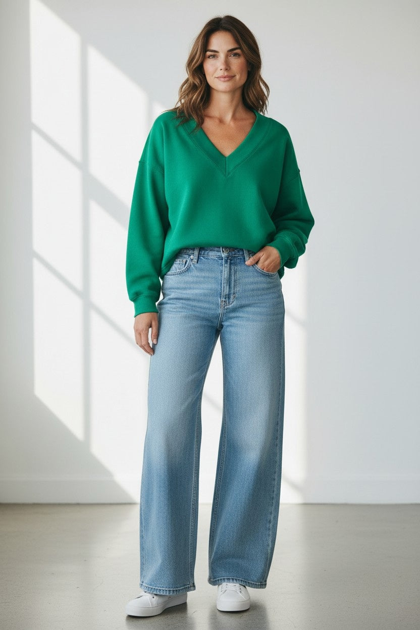 A woman stands indoors in the KMJ - TUCKER SWEATSHIRT by KMJ, a bright green relaxed fit V-neck, paired with light blue high-waisted wide-leg jeans and white sneakers. She smiles slightly with one hand in her pocket as sunlight streams in.
