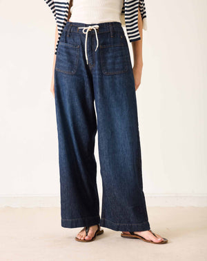 Wearing the MERSEA Londonberry Drawstring Jeans in Night Sky—a pair of wide-leg dark denim jeans with a comfortable drawstring waist—from MERSEA, styled with a cream sleeveless top, a striped shirt draped over the shoulders, and brown sandals, standing against a plain light background.