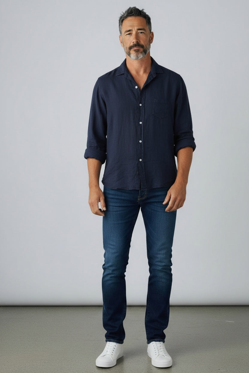 A man with short gray hair and a beard wears the FRANK &amp; EILEEN Finbar Men’s Button Up Shirt in Italian Linen Navy, paired with blue jeans and white sneakers. He stands on a gray floor against a light gray background, looking at the camera with a neutral expression.