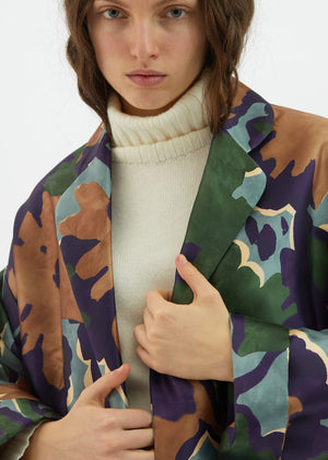 A person with long hair is sporting the ALYKI LABORATORIO PRINTED SATIN JACKET, featuring a green, purple, and brown floral pattern, over a white turtleneck sweater. With one hand in the pocket of their black pants, they perfectly blend an elegant color outfit against a plain white background.