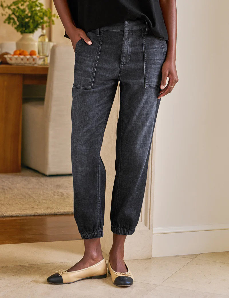 A person stands indoors with hands in the utility pockets of FRANK &amp; EILEEN’s Jameson Jogger Utility Jean in 1981 Wash. They pair it with a black top and tan ballet flats with black toe caps. A dining table and chairs are visible behind them.