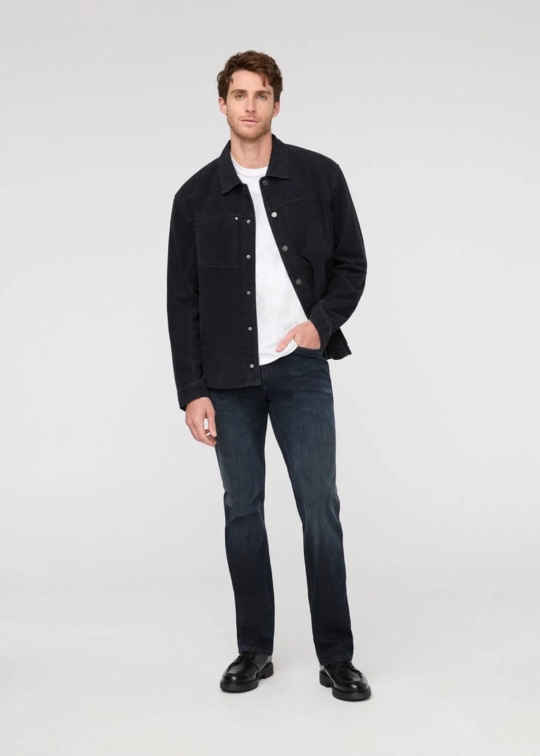 A man stands against a plain background wearing a black jacket, white t-shirt, DU/ER - PERFORMANCE DENIM+ SLIM STRIAGHT PANTS IN CARBON STONE, and black shoes, with one hand in his pocket and a neutral expression.