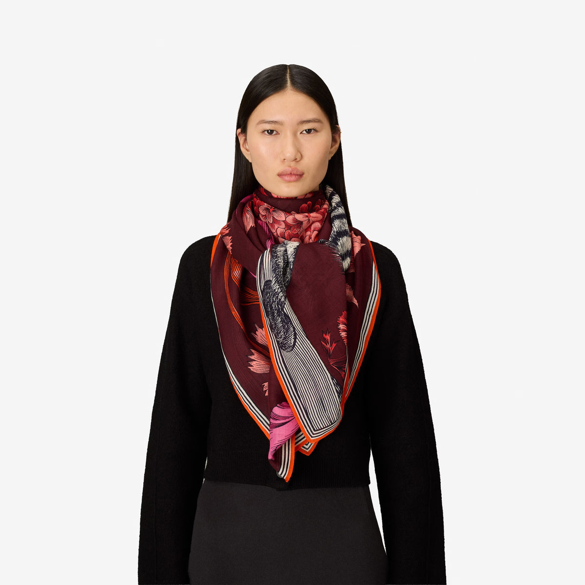 A woman with straight dark hair wears a black sweater and the INOUI EDITIONS - GUSTAV WOOL SCARF IN BURGUNDY by INOUI—a large (130 x 130 cm), multicolored patterned scarf crafted in India with cozy wool in rich red, pink, and burgundy tones—set against a plain white background.