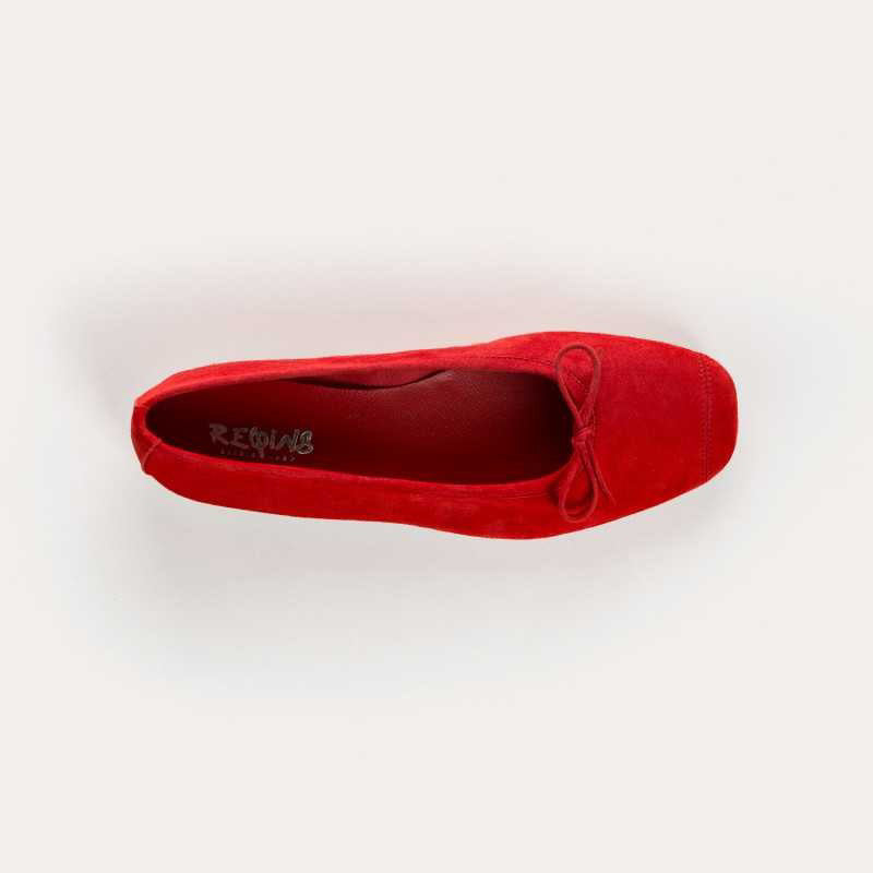 A person in white jeans, a beige coat, and carrying a round straw bag stands on light stone pavement, wearing REQINS HARMONY suede ballerina flats in rouge by REQINS.