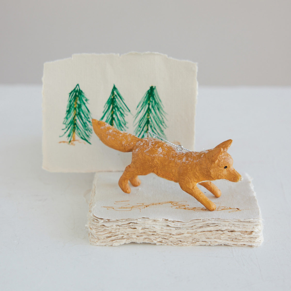 A small resin VINTAGE FOX ORNAMENT stands on a stack of textured paper in front of a woodland decor illustration featuring three green pine trees.