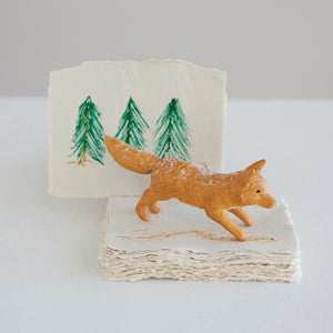 A small resin VINTAGE FOX ORNAMENT stands on a stack of textured paper in front of a woodland decor illustration featuring three green pine trees.