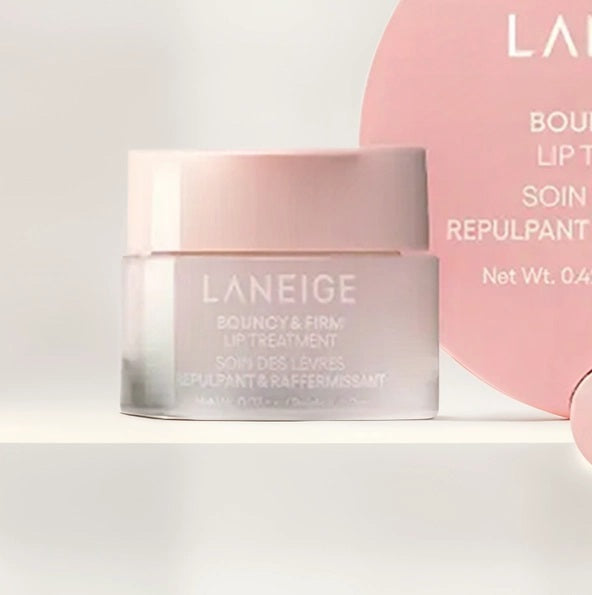 A pink LANEIGE - BOUNCY & FIRM LIP MASK (12g) by KOSMIC KBEAUTY SKINCARE sits open with its lid off, displaying the product inside. Beside the round container lies a small applicator. The label reads “Bouncy & Firm Lip Mask.” Designed to plump and moisturize dry lips overnight, this lip mask leaves your lips soft and healthy-looking.