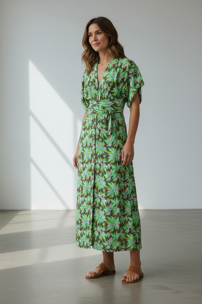 A woman stands indoors in the FRANCO FERRARI - JERSEY PRINT DRESS, a long green botanical wrap dress with short sleeves, paired with brown sandals. She has wavy brown hair and smiles softly as sunlight streams through the window.