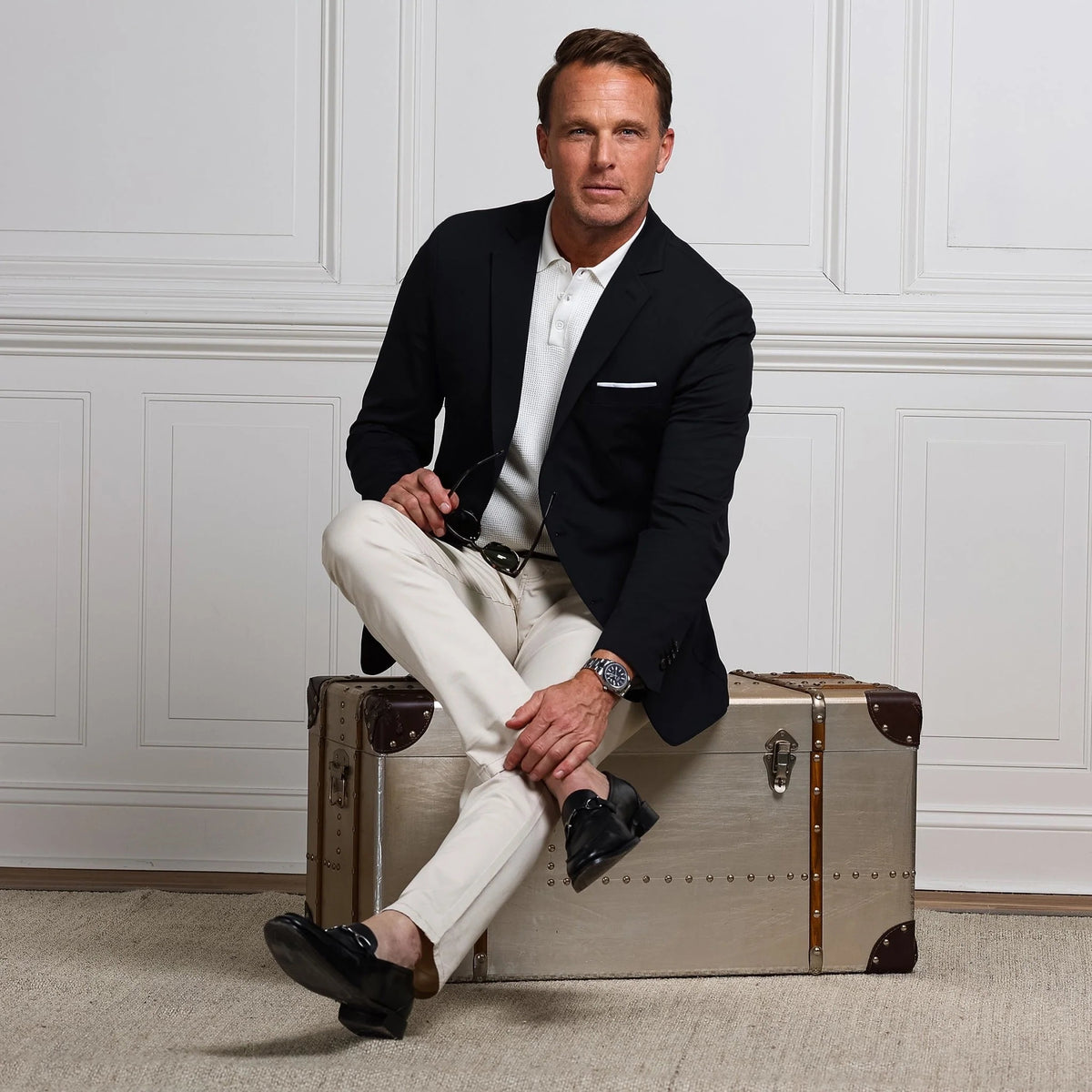 A man in the COLLARS AND CO Maverick Performance Blazer in Stealth Black, layered over a white t-shirt and beige pants, stands by a white paneled wall, looking to the side as he adjusts his blazer.