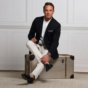 A man in the COLLARS AND CO Maverick Performance Blazer in Stealth Black, layered over a white t-shirt and beige pants, stands by a white paneled wall, looking to the side as he adjusts his blazer.