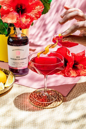 A transparent glass bottle holds a rich, dark red liquid labeled "YES COCKTAIL CO - Hibiscus Rose Cocktail Mixer." Handcrafted in California and available in a 16 fl oz (473ml) size, the white-and-red-toned label indicates its perfect suitability for creating cocktails or an exotic floral spritzer.