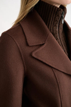 A close-up shows a person wearing the KINROSS Knit Insert Notch Collar Coat in Chocolate, featuring a wide lapel and crafted from luxurious wool cashmere, layered over a dark brown high-neck zip-up sweater. Only the neck, shoulder, and part of the face are visible.
