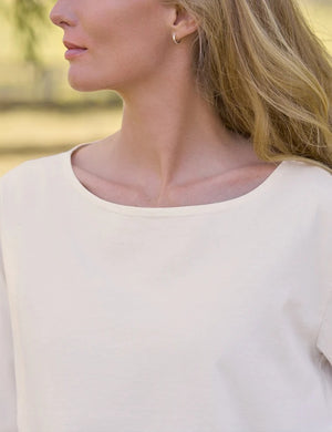 A woman with long blonde hair stands outdoors in front of a wooden fence, wearing the FRANK & EILEEN ELLA Long Sleeve Boatneck Tee in Vintage White and blue jeans, with trees and grassy fields in the background.