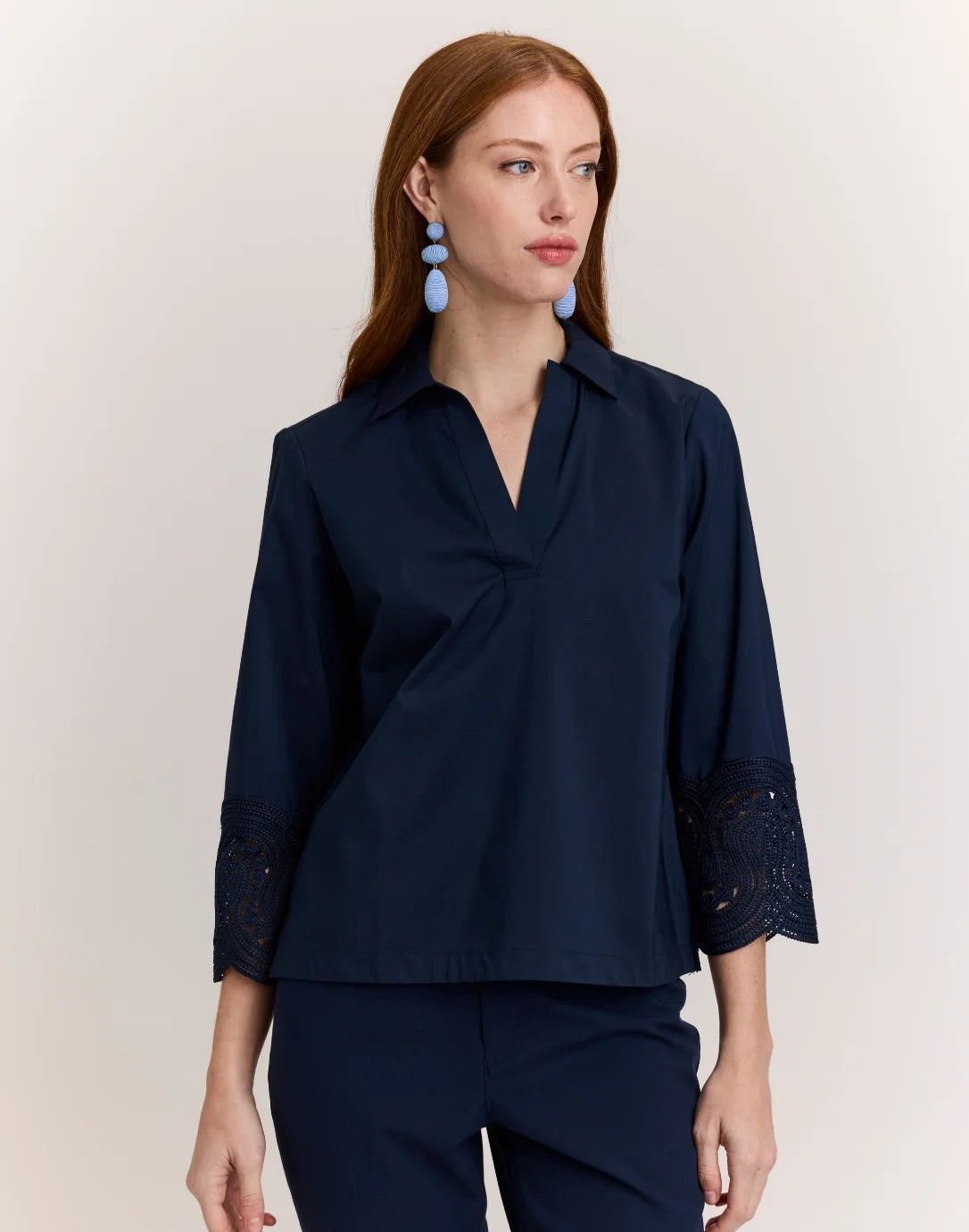 A woman with long red hair wears the HINSON WU MONA Bracelet Sleeve Stretch Polished Cotton Lace Top in navy—a comfortable, stretch cotton blouse featuring three-quarter sleeves with lace accents—paired with navy pants. She accessorizes with blue statement earrings and stands against a plain light background.