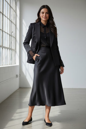 A woman stands against a plain light background wearing the MILVA MI - BLACK SATIN BLAZER by MILVA, paired with a matching long skirt, black blouse, black heels, and gold hoop earrings.