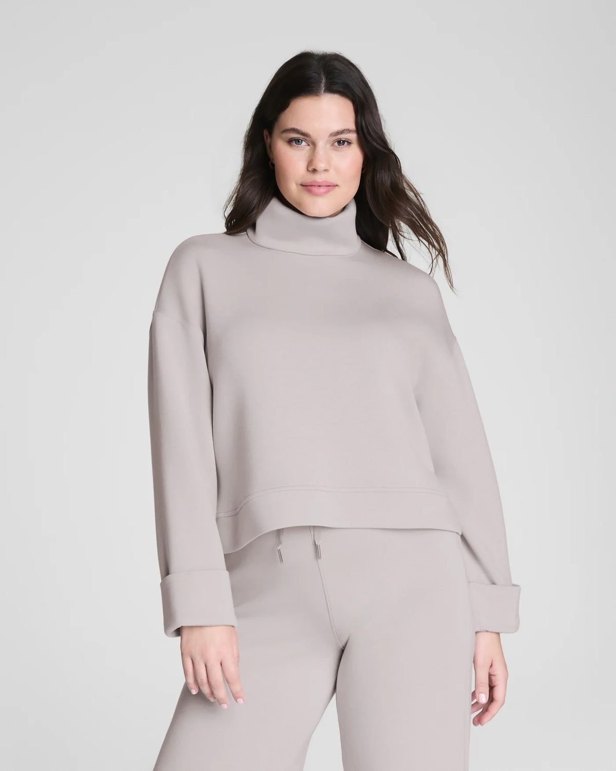 A woman confidently poses in the SPANX - AIRESSENTIALS TURTLENECK TOP and matching pants, crafted from ultra-drapey spacer fabric, against a plain light gray background with a slight smile.