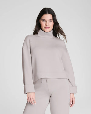 A woman confidently poses in the SPANX - AIRESSENTIALS TURTLENECK TOP and matching pants, crafted from ultra-drapey spacer fabric, against a plain light gray background with a slight smile.