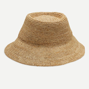A woman with long wavy blonde hair, in a sleeveless white top, wears the WYETH HATS - TALI RAFIA BUCKET HAT IN NATURAL by WYETH, shown in profile against a plain light background.