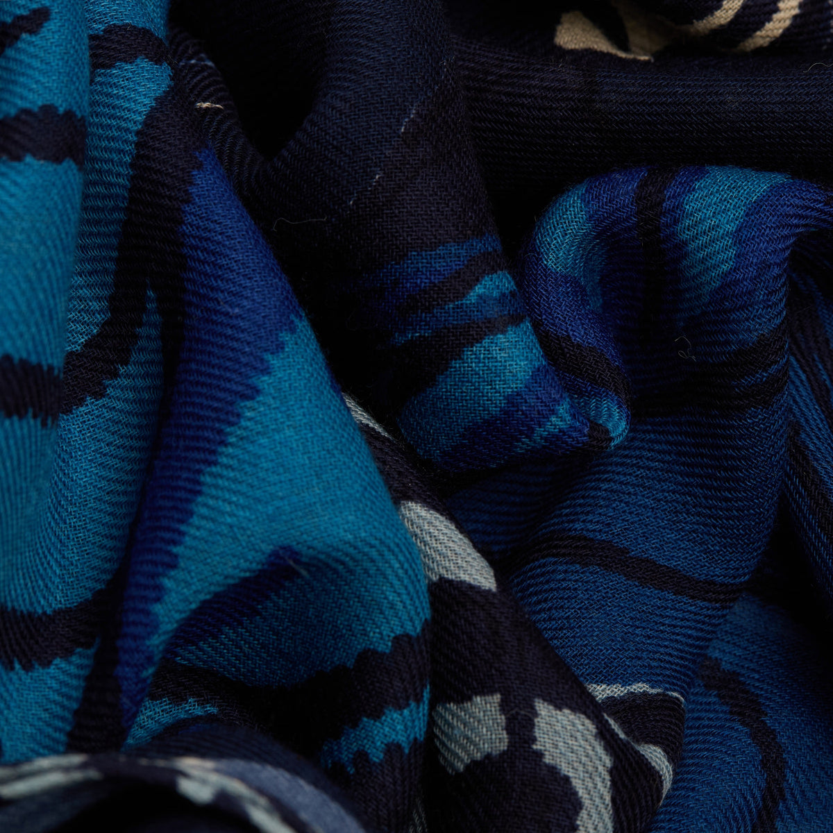 A woman with light hair pulled back wears a light gray jacket and the INOUI EDITIONS PAPILLON (BUTTERFLIES) square wool scarf in blue by INOUI. She stands against a plain white background, looking straight at the camera with her hands in her pockets.