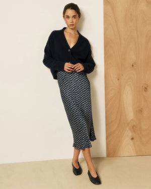 A woman stands in a bright room wearing the HARTFORD - JIMO Bias Cut Viscose Skirt in Navy with White Dot, paired with a navy cardigan and black flats. She has wavy brown hair and is smiling softly as natural light streams through large windows.