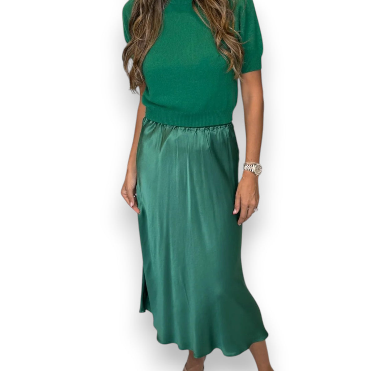 A woman models the ROSE &amp; CROWN - SATIN MIDI SKIRT in silky blue, paired with a short-sleeved light blue knit top, standing against a white background. Her head is not visible in the image.