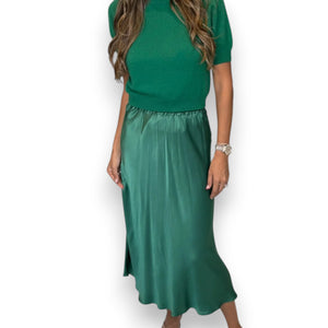 A woman models the ROSE & CROWN - SATIN MIDI SKIRT in silky blue, paired with a short-sleeved light blue knit top, standing against a white background. Her head is not visible in the image.