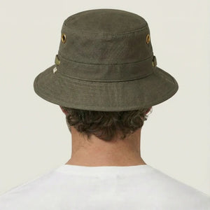 A person wearing the TILLEY - ICONIC T1 BUCKET HAT IN OLIVE with yellow eyelets and a white crew-neck shirt looks slightly to the side against a plain light background. The TILLEY hat offers UPF 50+ sun protection.