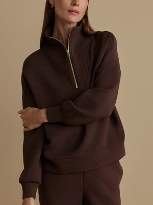 A person wears an oversized, dark brown VARLEY - HAWLEY HALF-ZIP SWEATSHIRT IN COFFEE BEAN MARL with a gold zipper.