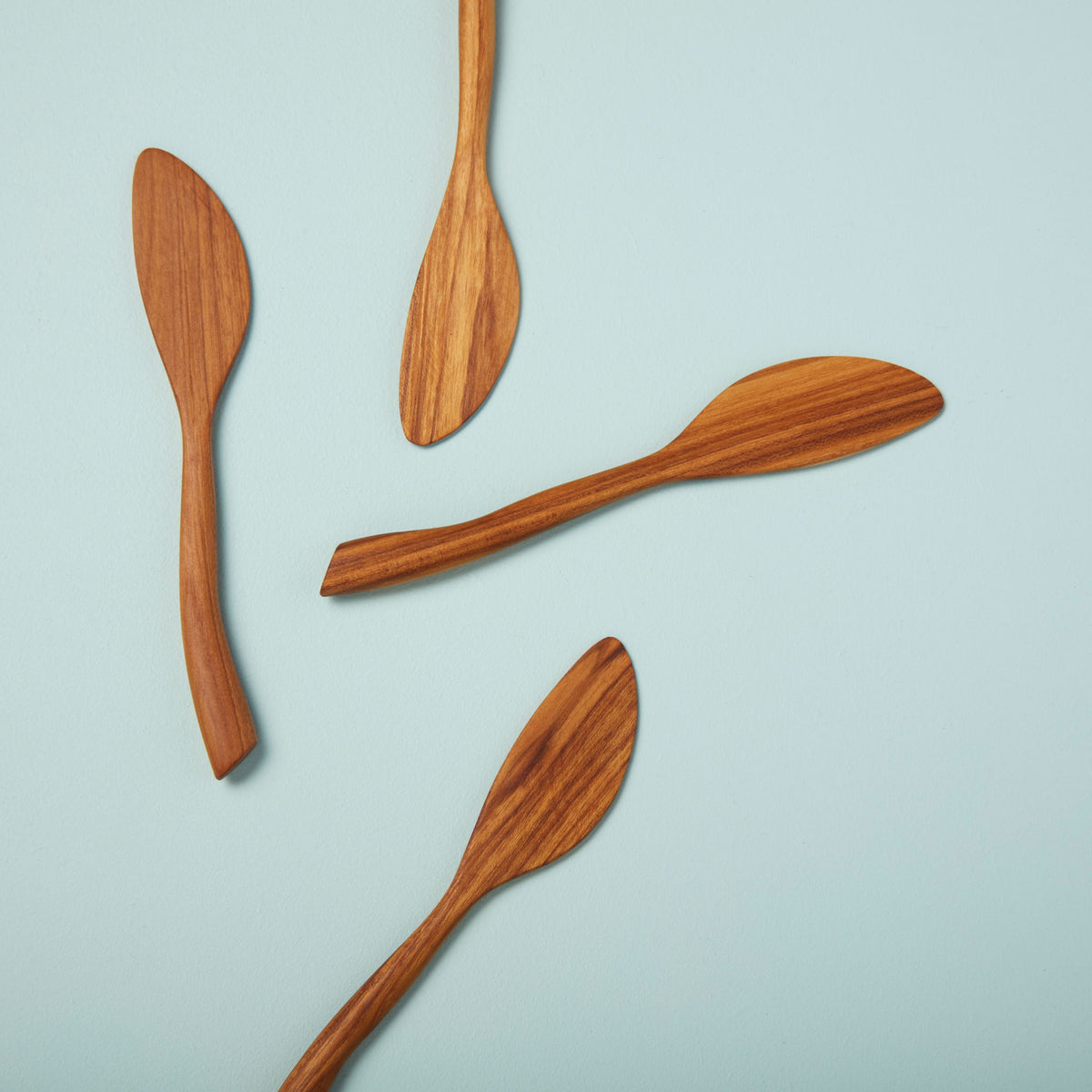 Four BE HOME, INC BEHOME - TEAK WAVE SPREADER knives with smooth, curved teak handles are arranged diagonally on a light blue surface, each blade facing a different direction.