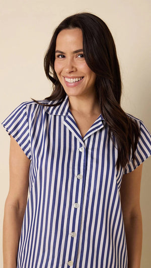 A person, smiling in a CAT'S PAJAMAS Classic Stripe Luxe Pima Short Set—a blue and white striped short-sleeve shirt with matching shorts—stands against a plain light beige background.
