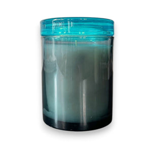 The MERSEA - SEA CHANGE JOLIE JAR CANDLE by MERSEA features white tea and sea salt-scented white wax in a cylindrical glass jar with a translucent blue lid, displayed against a white background.