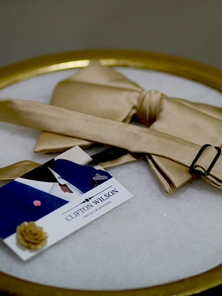 A gold bow tie with an adjustable strap rests on a round surface with a gold edge, next to a business card featuring a blue suit and the name 