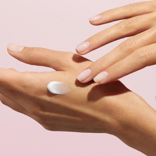 With perfectly manicured nails, a person applies MANUCURIST - COSMOS ORGANIC HAND CREAM, ROSE NOUVELLE from MANUCURIST INC to the back of their hand against a soft pink background, emphasizing the importance of caring for hands and nails.