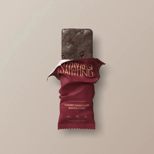 A maroon protein bar wrapper labeled “STARS+HONEY” displays the flavor “CHERRY CHOCOLATE WAFFLE CONE” with nutrition highlights: 8g collagen, 15g protein, and 2g sugar, all set against a beige background. This indulgent treat puts the rich chocolate and tart cherry combination front and center.