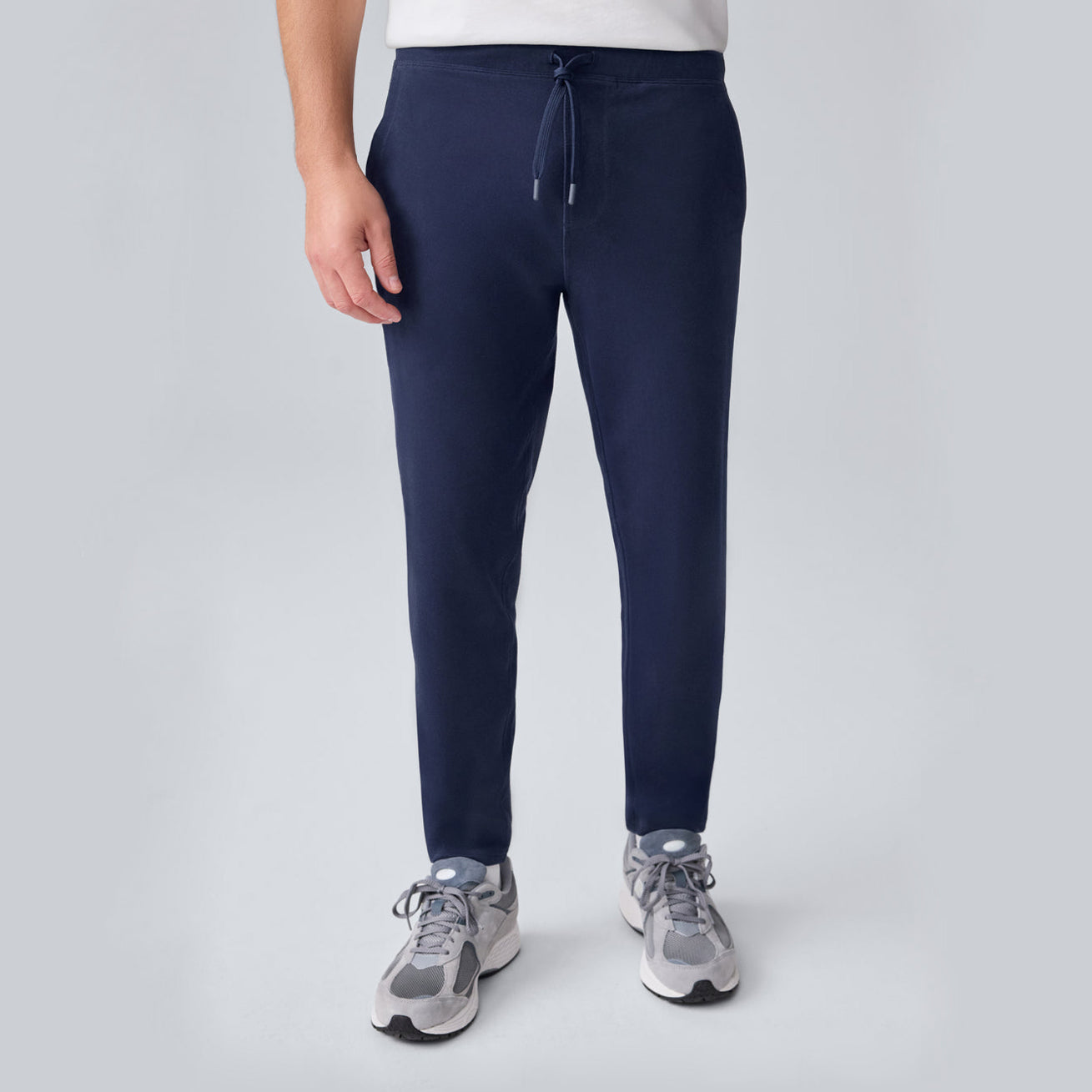 A person wearing a white shirt, navy blue jogger pants with a drawstring, and gray sneakers stands against a plain light gray background. The person's face is not visible.