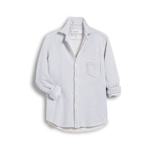 A woman smiles in a bright kitchen, wearing the FRANK & EILEEN EILEEN Relaxed Button Up Sweatshirt in Grey Melange. She pairs it with blue wide-leg jeans and tan boat shoes, leaning against a red table with one hand in her pocket.