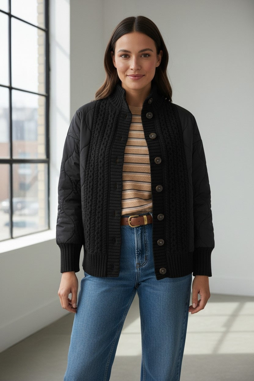 A woman stands indoors by large windows, wearing a striped sweater, blue jeans, and the KINROSS CASHMERE Quilted Cable Cardigan in black from KINROSS. She has straight, dark hair and is smiling gently at the camera.