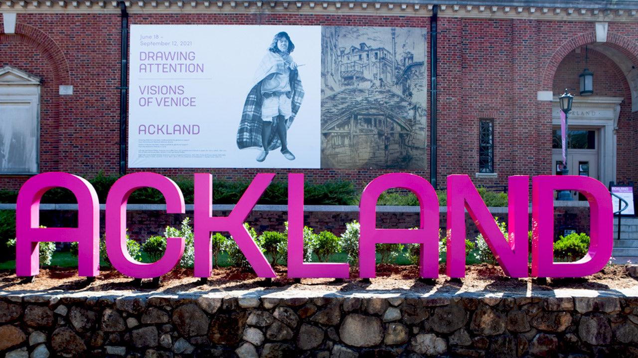 Large pink letters spell “ACKLAND” in front of a brick building with arched doorways. A banner advertises “Drawing Attention: Visions of Venice” at the Ackland Art Museum. Shrubs and stone wall in foreground.
