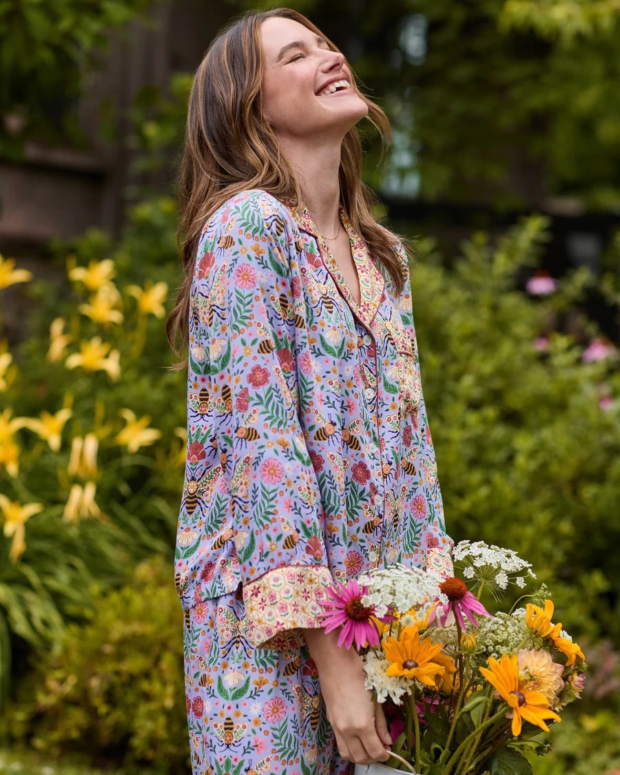 A person wearing the PRINTFRESH - BEE TAPESTRY ECO SATIN SOHO SET IN LAVENDER, featuring vintage-inspired bee and floral vine prints, holding a flower behind their back in a garden.