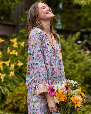 A person wearing the PRINTFRESH - BEE TAPESTRY ECO SATIN SOHO SET IN LAVENDER, featuring vintage-inspired bee and floral vine prints, holding a flower behind their back in a garden.