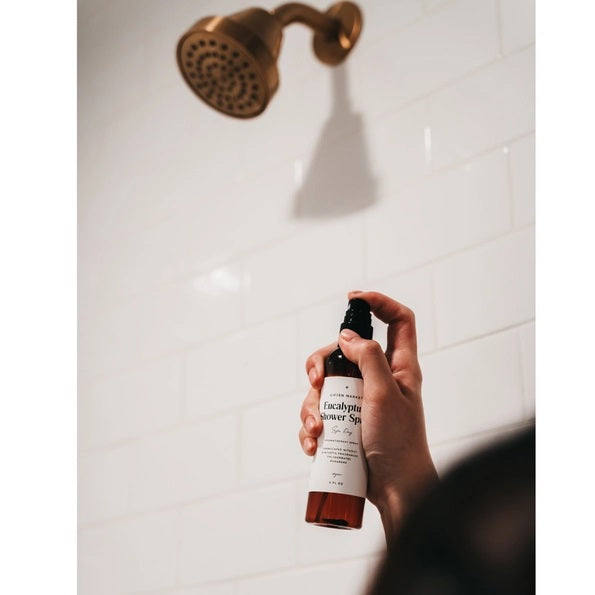 A hand holds a brown spray bottle labeled “SPA DAY EUCALYPTUS SHOWER SPRAY” by GIFTEN MARKET—an uplifting eucalyptus aromatherapy mist—set against a black showerhead and white marble shower wall, ideal for transforming your shower into a spa-like escape at home.