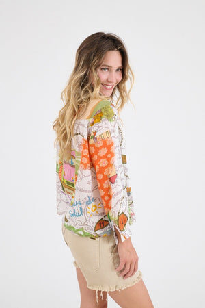 A woman with wavy blonde hair exudes casual elegance in the STORIATIPIC SUZIE GABIE BLOUSE, a long-sleeve cotton shirt with a colorful map print, paired with beige frayed shorts. She smiles softly against a plain white background, one hand in her pocket.