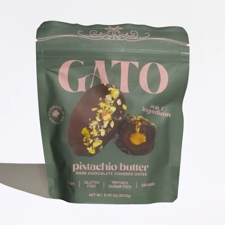 A green package of GATO DATES - CHOCOLATE COVERED PISTACHIO BUTTER DATES by GATO DATES, featuring premium Medjool dates coated in dark chocolate. The image displays the treat split open, revealing a creamy pistachio butter filling and a sprinkling of pistachios on top. Packaging text highlights that it is vegan, plant-based, gluten-free, and organic—offering a delightful chocolate experience from Dubai.