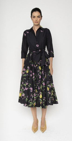 A woman stands against a plain backdrop wearing the LAVI COUTURE Spritz Black Floral Dress, featuring vibrant pink and green patterns on a citrus and floral print mid-length skirt with long sleeves, paired with beige pointed high heels.