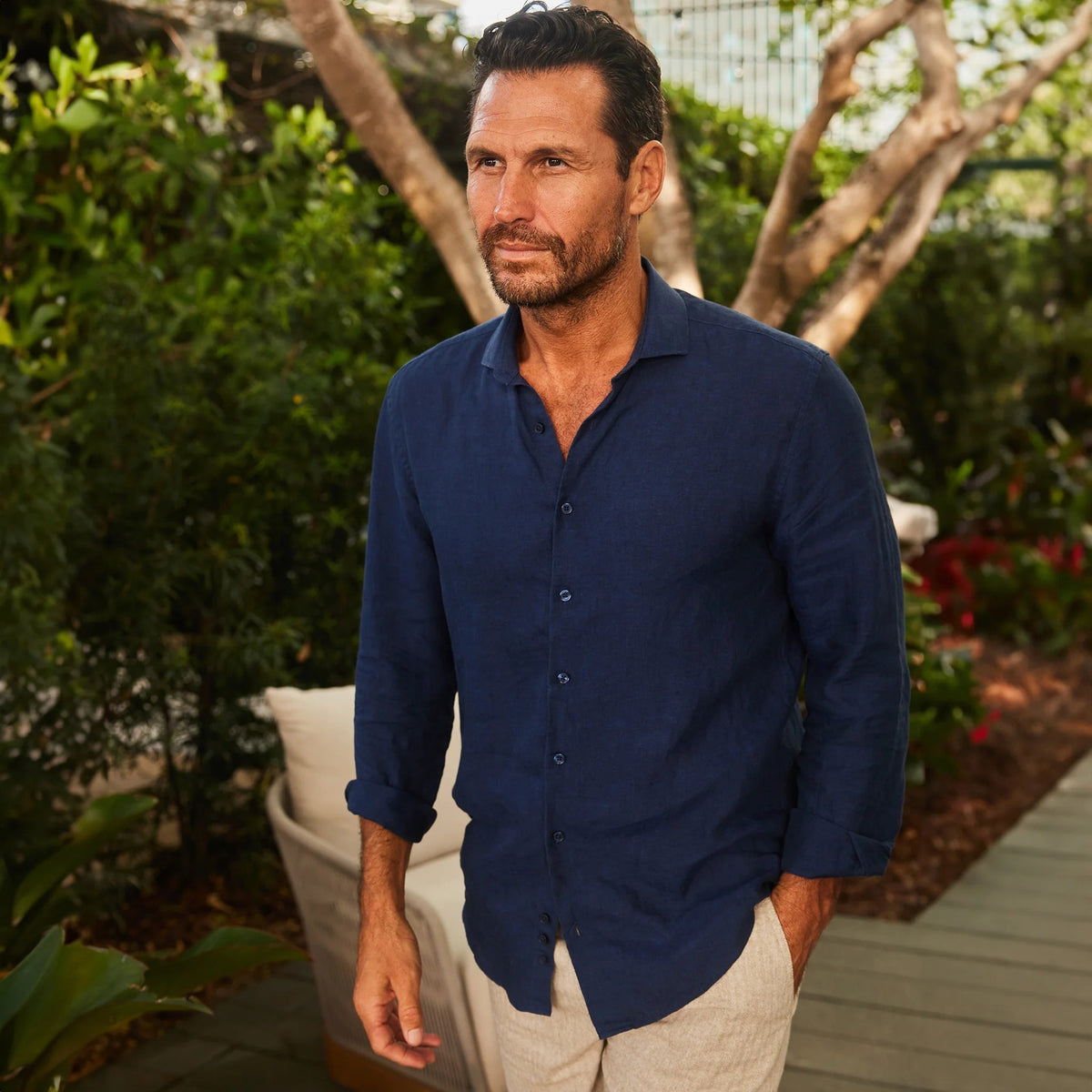 A man with short dark hair and a beard, wearing the COLLARS AND CO Toscano Linen Shirt in Navy with a reinforced collar, sits on an outdoor sofa against green foliage, looking at the camera with a neutral expression.