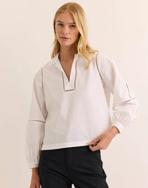 A woman with long blonde hair wears the HINSON WU LAURA Long Sleeve Lace Trim Top in white, crafted from stretch polished cotton and featuring decorative cut-out lace details along the sleeves and neckline. She stands against a neutral background, looking toward the camera with a slight smile. This stylish blouse by HINSON WU is also machine washable, offering effortless care alongside its sophisticated design.
