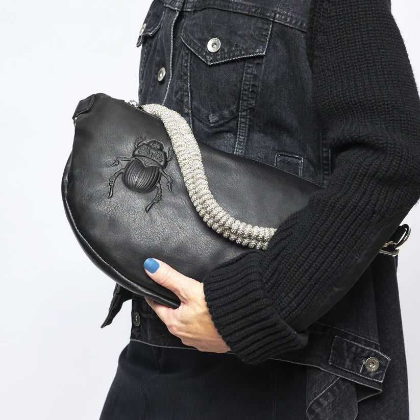 The GANRIELE FRANTZEN - SCARAB LEATHER XL BELT BAG IN BLACK WITH EXTRA CRYSTAL STRAP by GABRIELE FRANTZEN is a black leather, crescent-shaped crossbody bag featuring a long black strap and an extra decorative crystal handle, displayed against a plain white background.