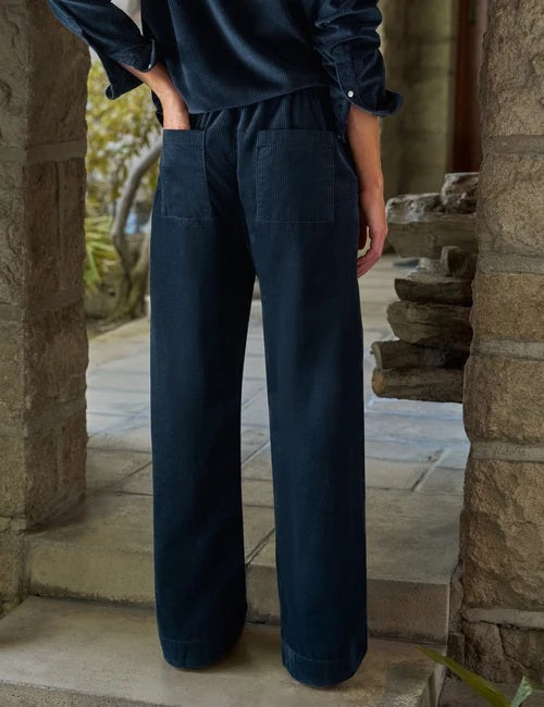A person stands indoors by a window, dressed in the FRANK & EILEEN MADELEINE Patch Pocket Pull On Pant in Italian Corduroy Classic Navy and a coordinating dark navy button-up shirt, paired with brown shoes. With one hand tucked into their pocket, they smile softly as sunlight streams into the minimalist room.