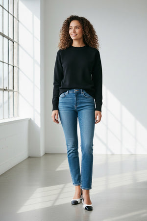A person stands against a plain backdrop, confidently showcasing their style in SPANX's KICK FLARE JEAN, made with 4-way stretch denim. They pair it with a black sweater and black-and-white heels, their long dark hair cascading as they pose with one hand in their pocket.